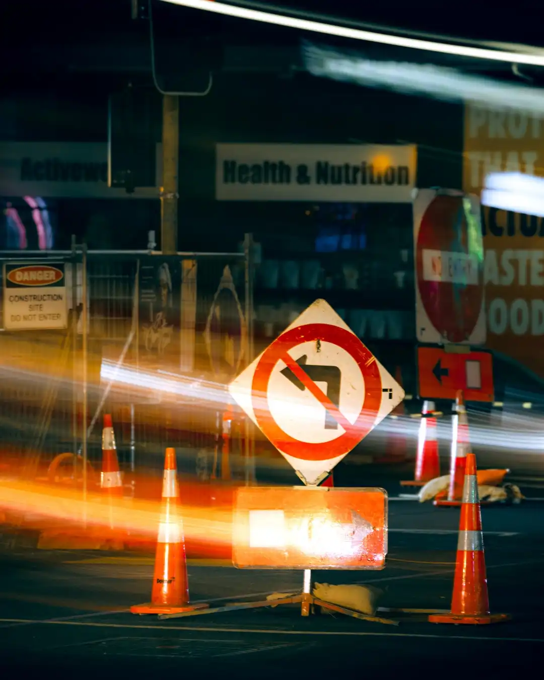 Construction sign with orange cones and traffic light trails.