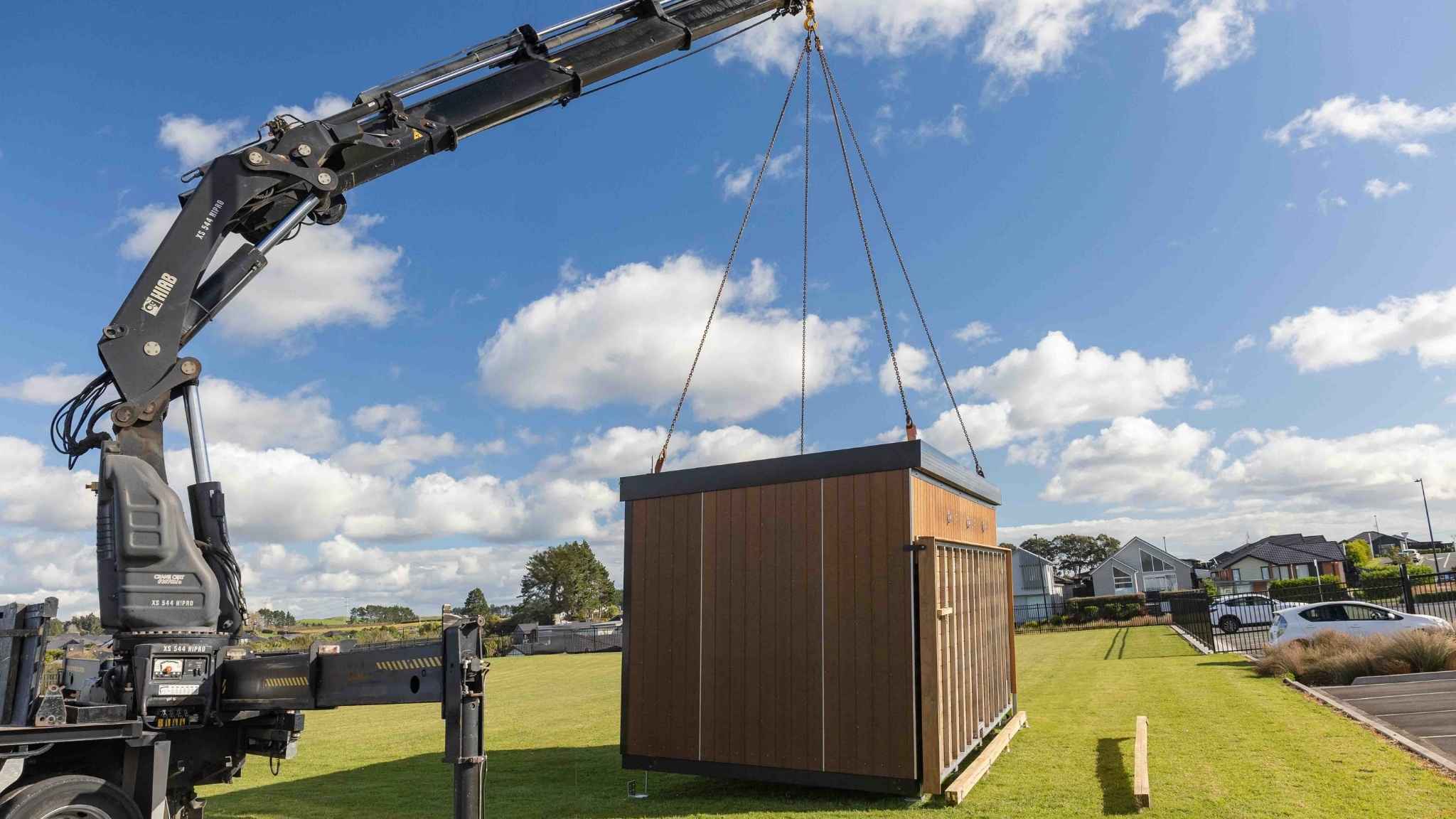 Crane lifting a small prefabricated cabin into place against blue sky and suburban backdrop.