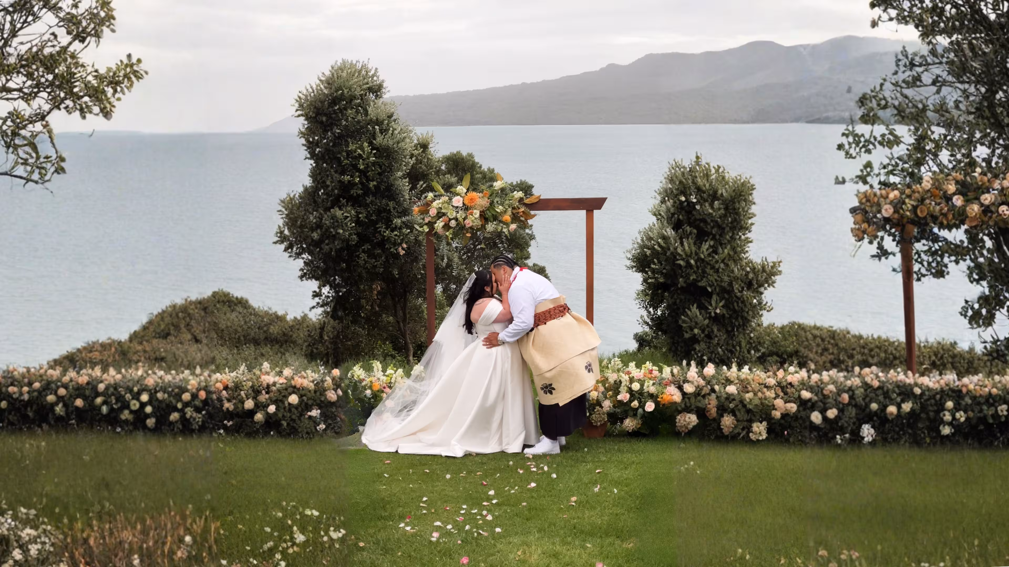 Bride and groom kissing under a floral arch overlooking the ocean at The Officers Mess Auckland wedding venue