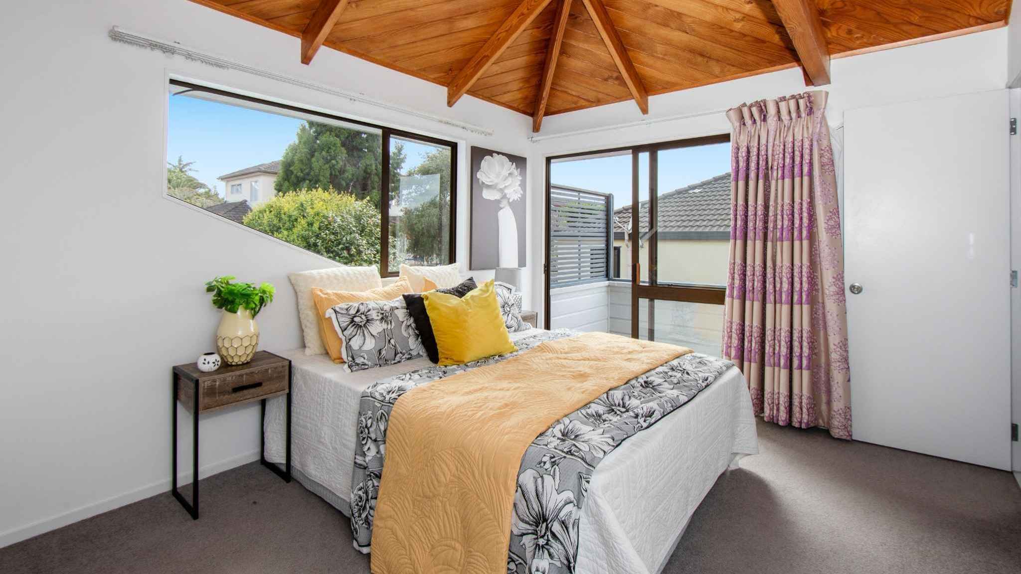 Sunlit bedroom with large corner windows, vaulted wooden ceiling, and stylish staging with yellow accents.