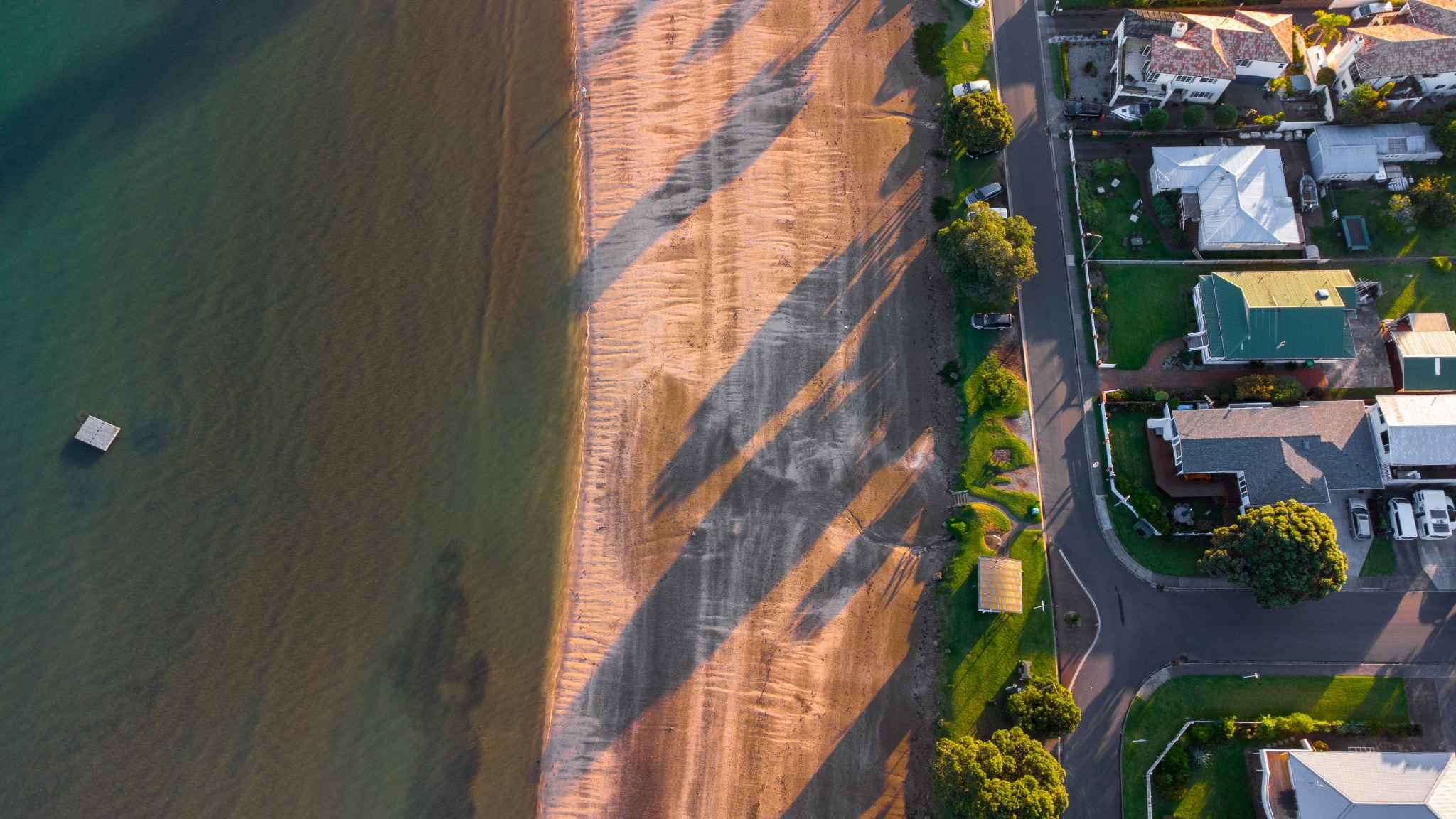Aerial drone image of golden sandy beach with long shadows at sunset, highlighting nearby coastal neighborhood of Arkles Bay.