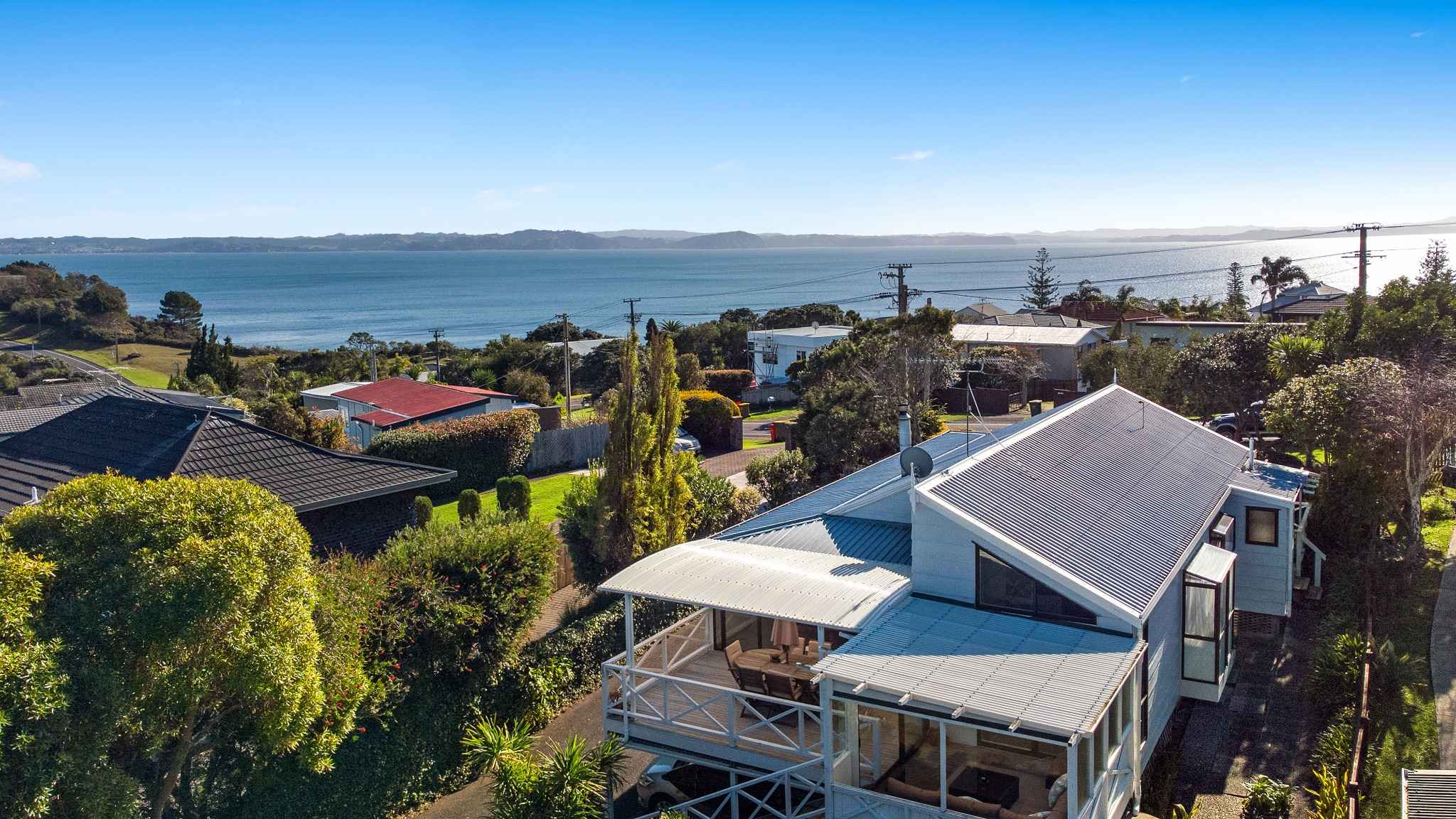 Aerial view of coastal home with blue roof and ocean backdrop in Whangaparaoa, Auckland real estate property for sale.